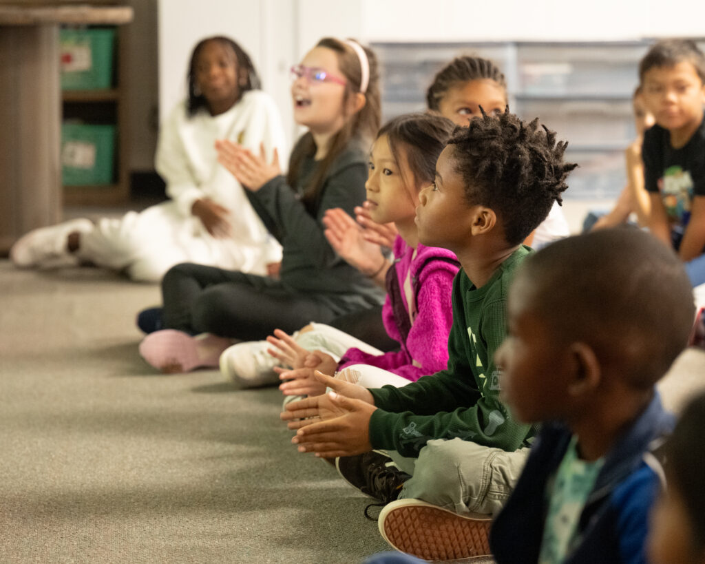 Students sit on the floor and engage in a morning meeting routine.