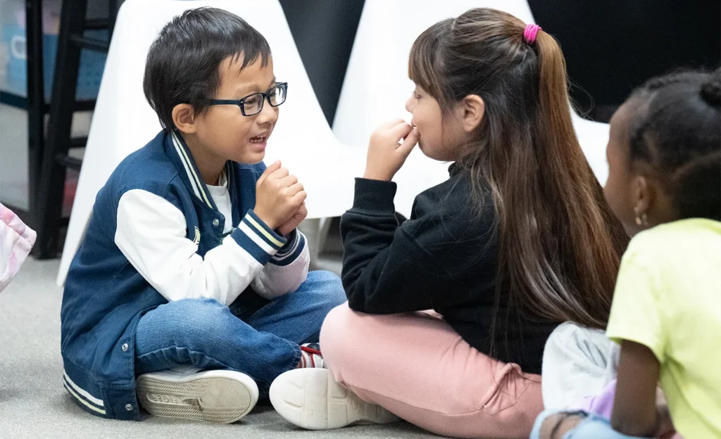 Two students sit on the floor of the classroom greeting one another during Morning Meeting with smiles on their faces.